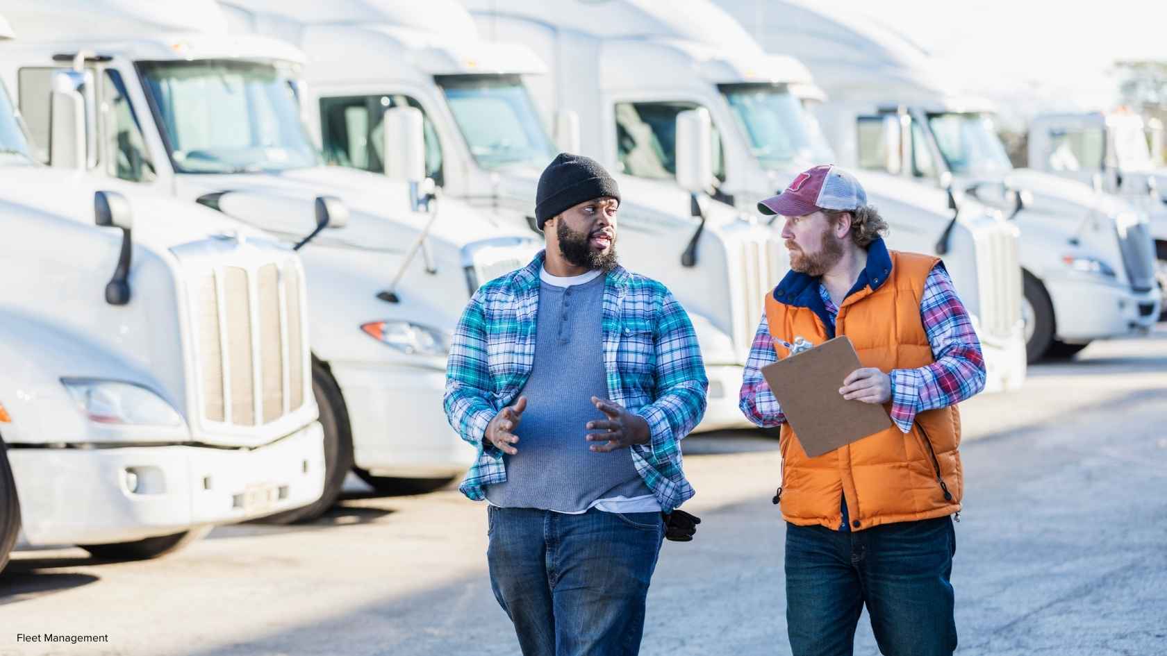 Two fleet managers discussing in front of a row of parked white semi-trucks, one holding a clipboard, in a fleet management setting.